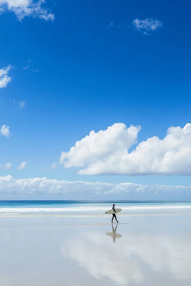 Surfer walking across a wide reflective beach under blue sky